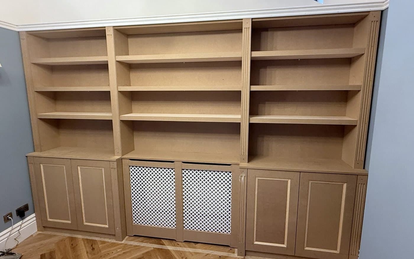 The finished bookcase with a radiator cover and panelled cupboard doors, fitted wall to wall in a living room