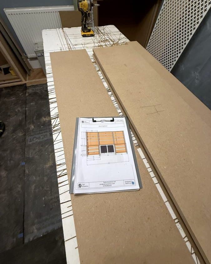 The printed drawing of a fitted bookcase on a clipboard, lying on cut MDF boards on the bench
